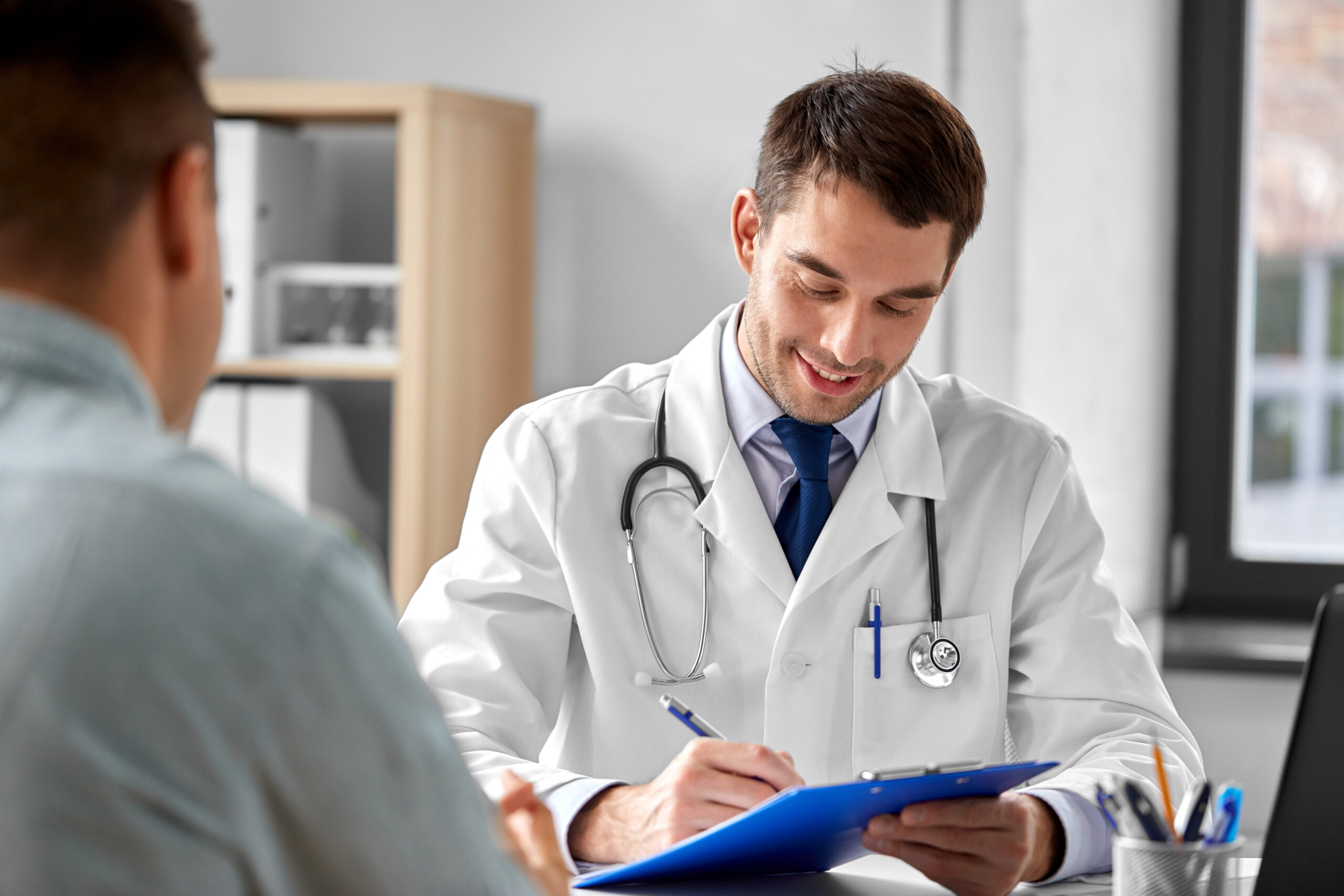 medicine, healthcare and people concept - doctor with clipboard talking to male patient at medical office in hospital