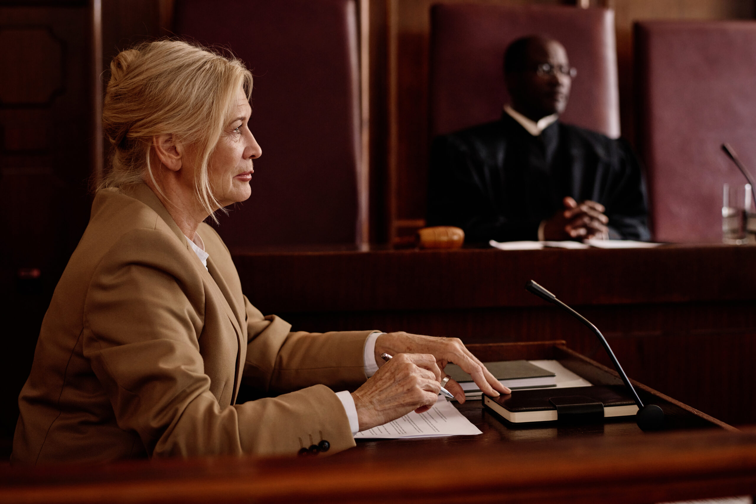 Side view of mature blond female witness or lawyer speaking in microphone while sitting against judge